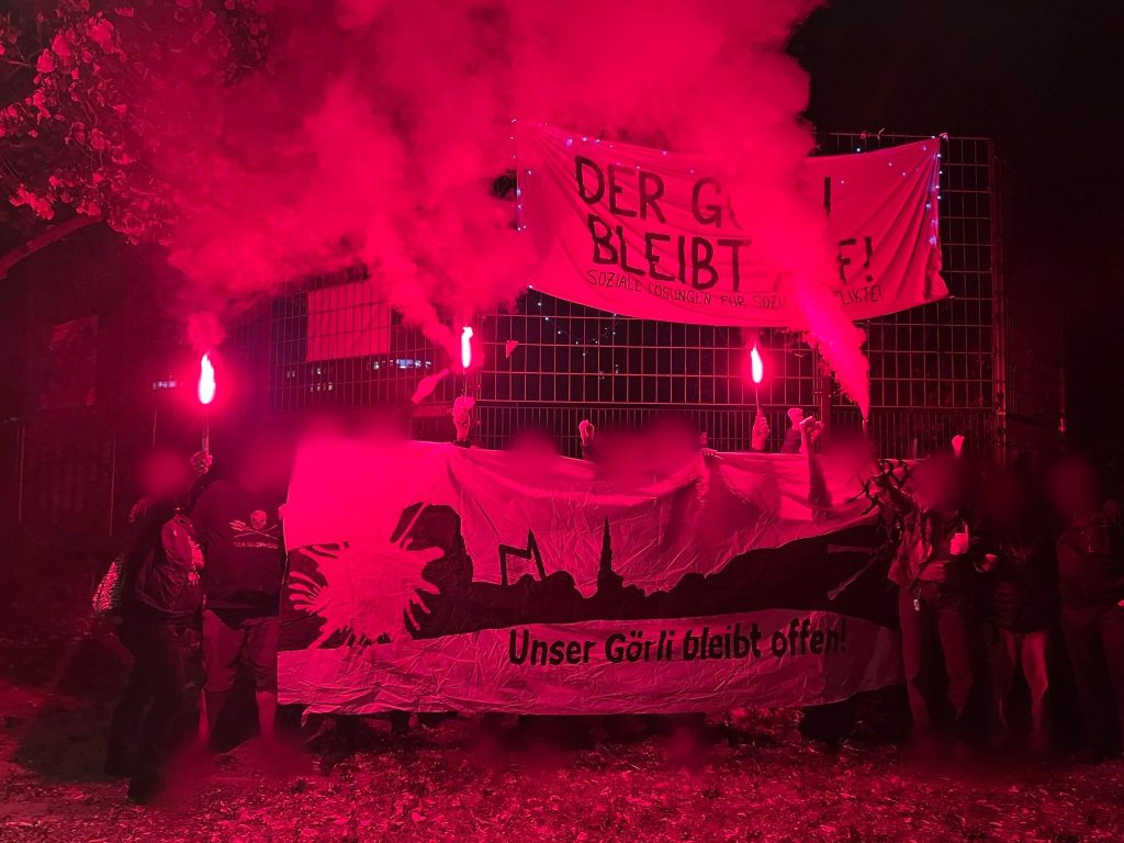Gruppe aktivistischer Menschen bei Nacht mit roter Pyro. Auf den Transpis steht: Unser Görli bleibt offen!